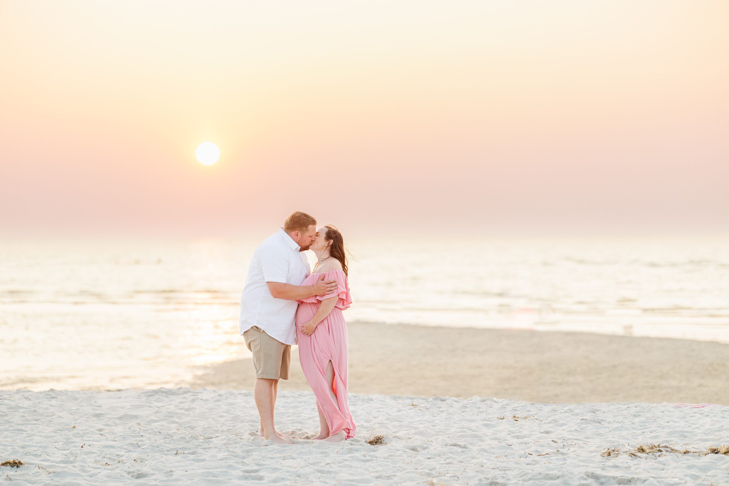 Couple kissing on the beach at sunset, the woman in a pink dress cradling her pregnant belly.