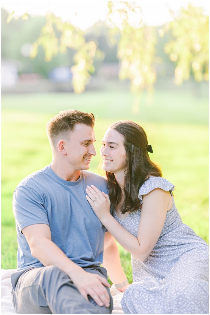 A couple sits on a blanket in the grass, smiling at each other. The woman’s hand, with an engagement ring, rests on the man’s chest.