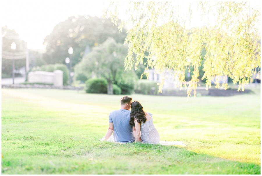 A couple sits on the grass under a tree, leaning toward each other as they look out over a sunlit field during an anniversary shoot with by a  Massachusetts photographer