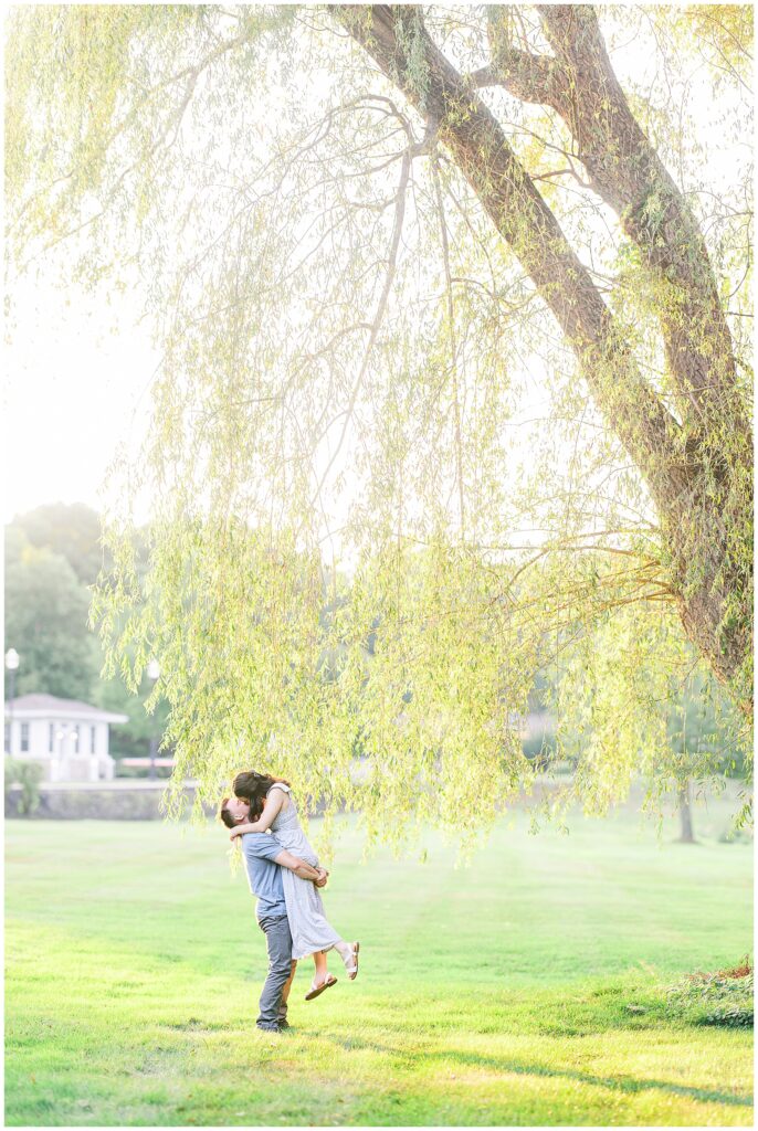 A couple kisses under a sunlit tree, with the man lifting the woman off the ground in a grassy field.