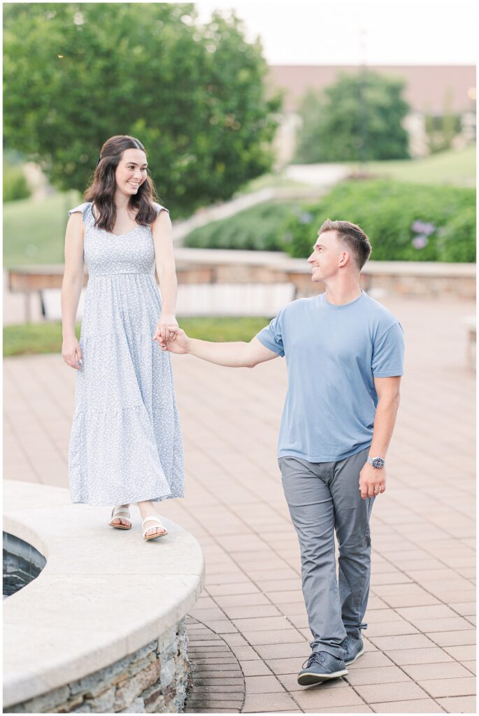 A couple walks hand in hand near a fountain, the woman leading while smiling down at the man. Greenery is visible in the background.