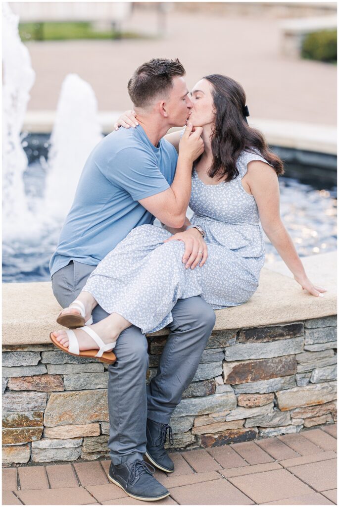 A couple sits by a fountain, kissing. The man has an arm around the woman, who gently holds his chin during a photoshoot with a Massachusetts photographer.