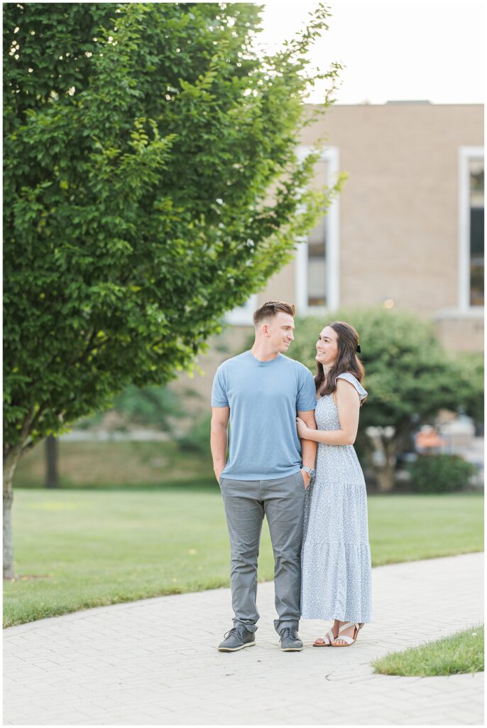 A couple poses on a path, with the man embracing. Trees and grass are in the background.