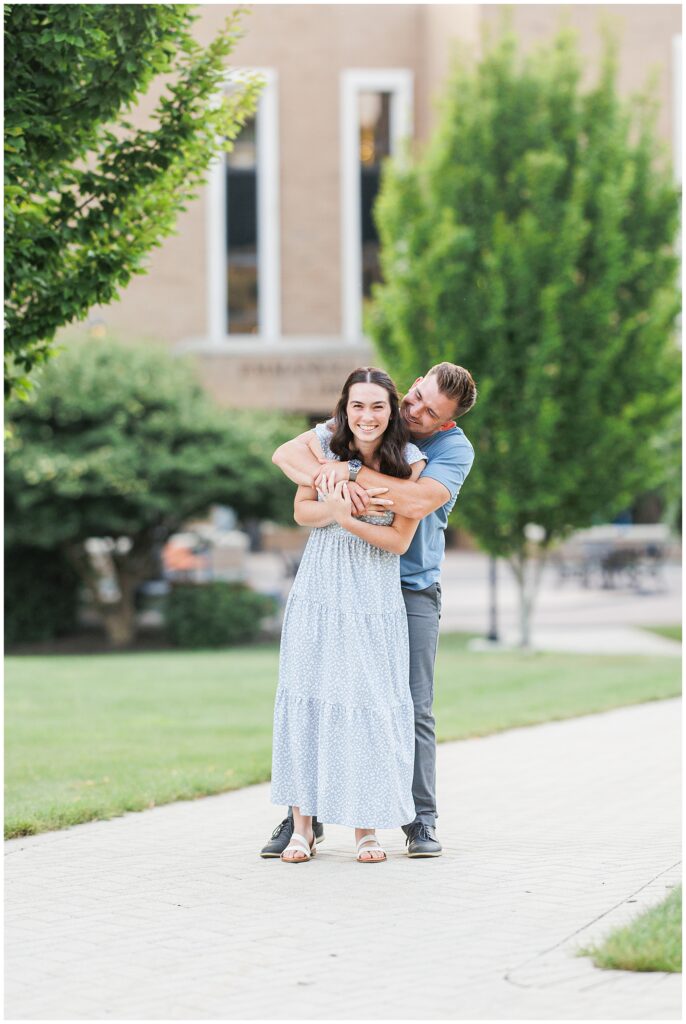 A couple poses on a path, with the man embracing the smiling woman from behind. Trees and grass are in the background.