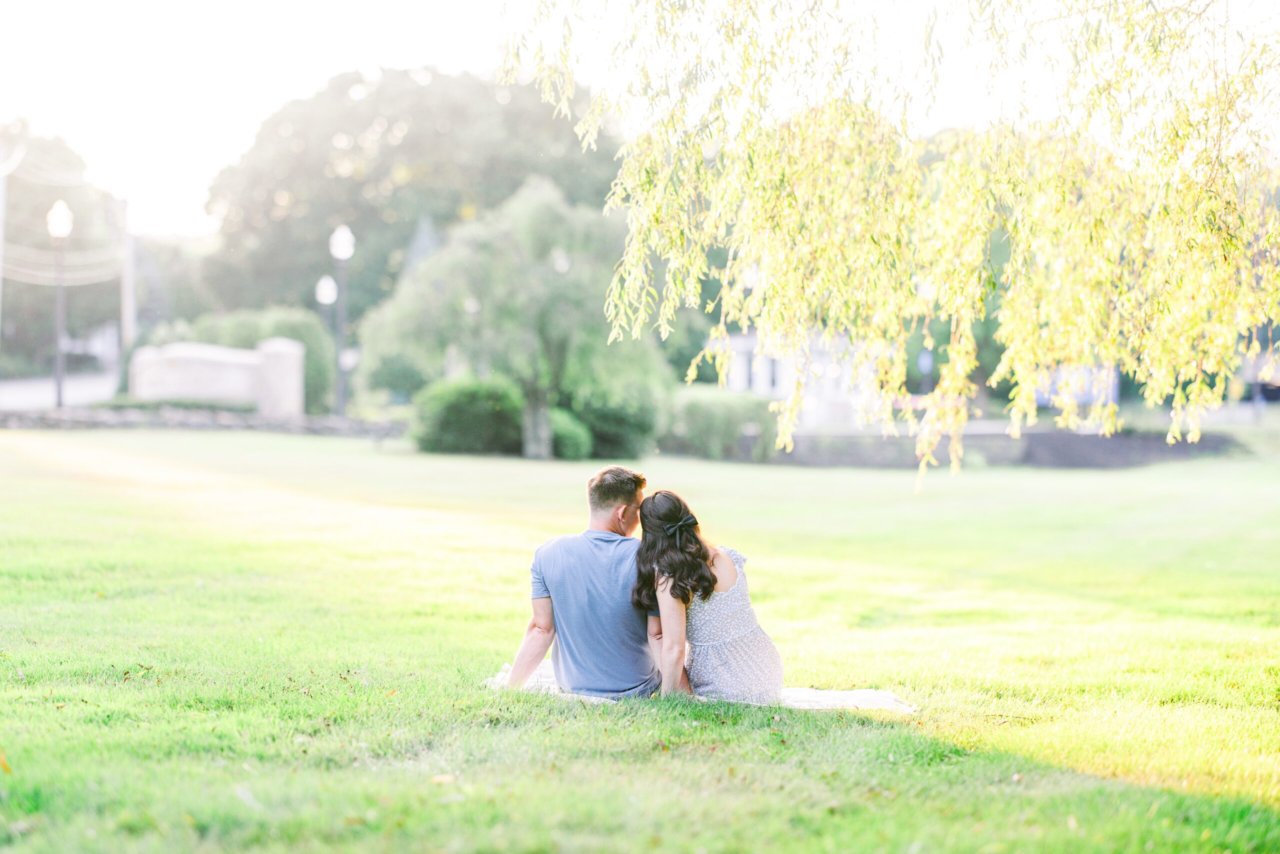 A couple sits on the grass under a tree, leaning toward each other as they look out over a sunlit field, taken by a Massachusetts photographer.