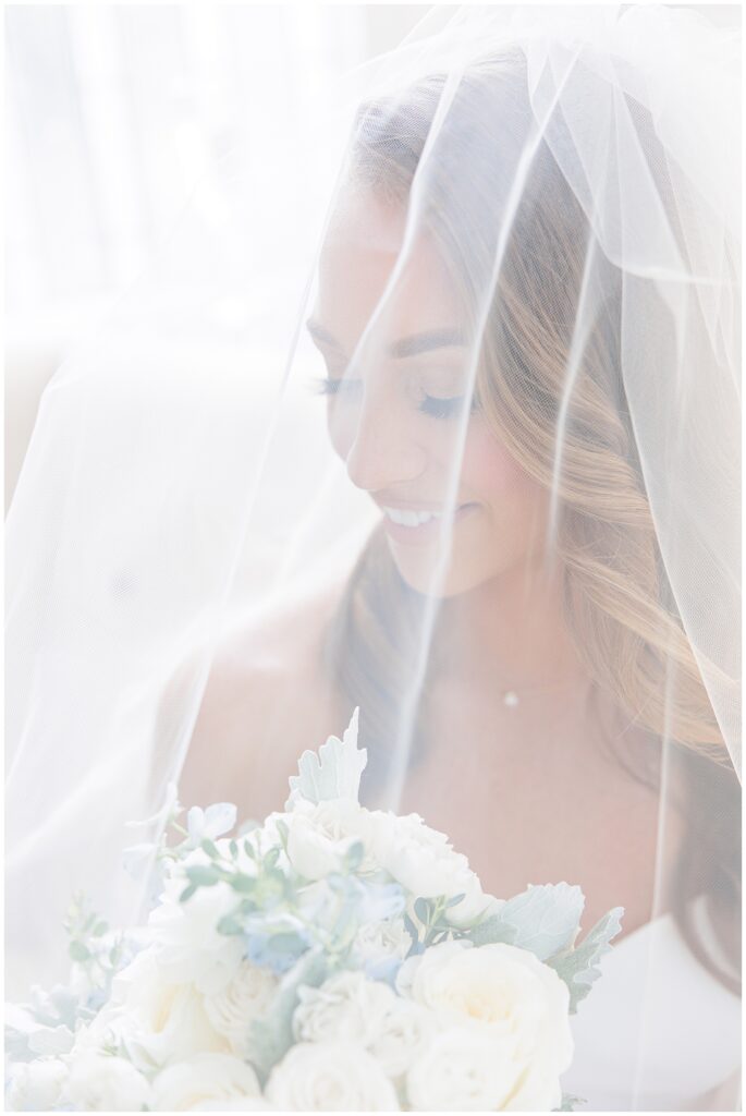 Bride smiling, holding a bouquet of white flowers, with a sheer veil covering her face.