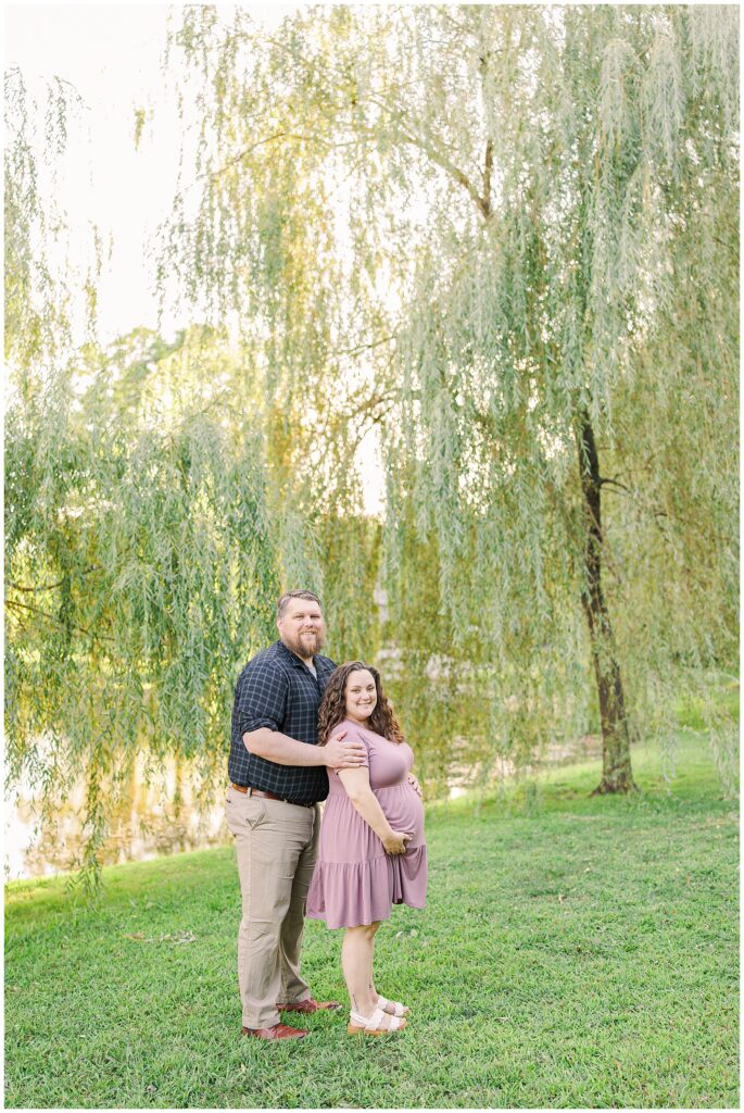 A couple standing together under willow trees by a pond at Larz Anderson Park. The woman is pregnant and wearing a lavender dress, while the man stands behind her, placing his hand on her belly.