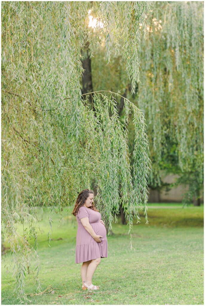 A pregnant woman in a lavender dress standing under hanging willow branches at Larz Anderson Park, smiling down at her belly.