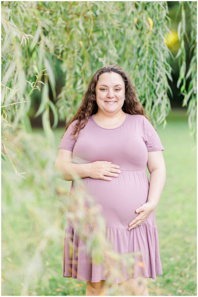 A portrait of a smiling pregnant woman in a lavender dress standing beneath willow tree branches at Larz Anderson Park. Her hands rest on her belly.