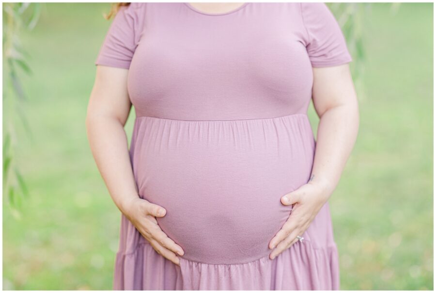 Close-up of a pregnant woman’s belly, with her hands gently cradling it. She is wearing a lavender dress, and the background is blurred greenery.