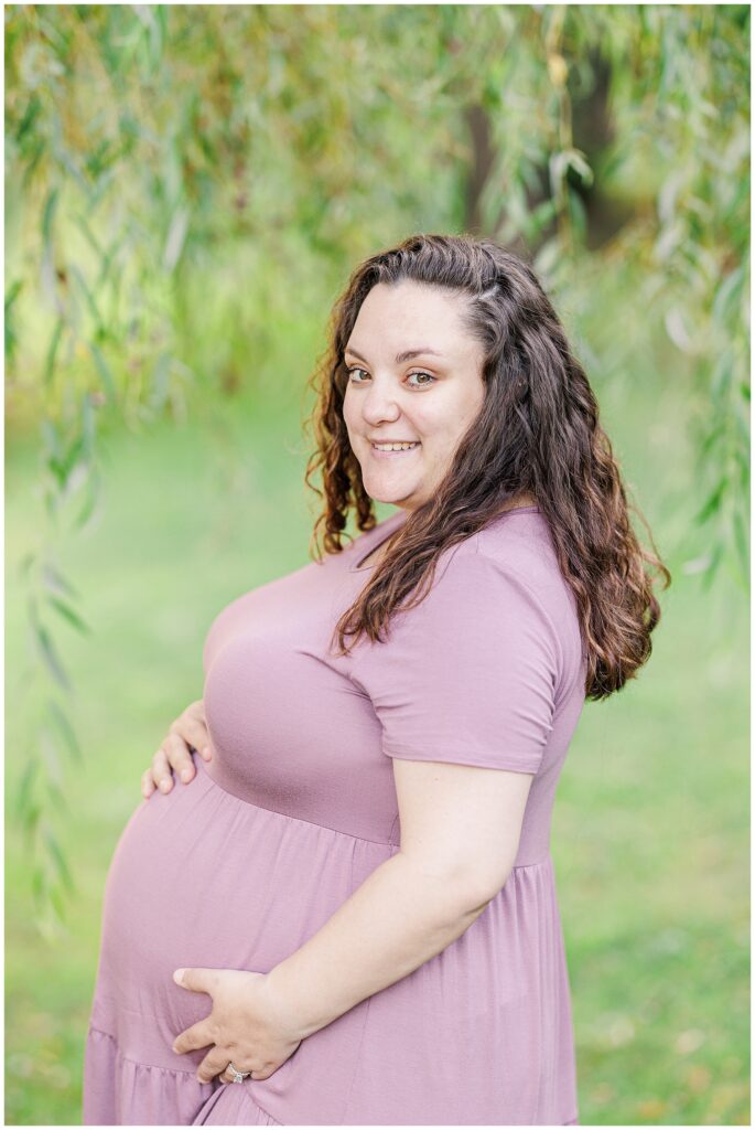 Side profile of a pregnant woman in a lavender dress standing under hanging willow branches at Larz Anderson Park, smiling as she cradles her belly.