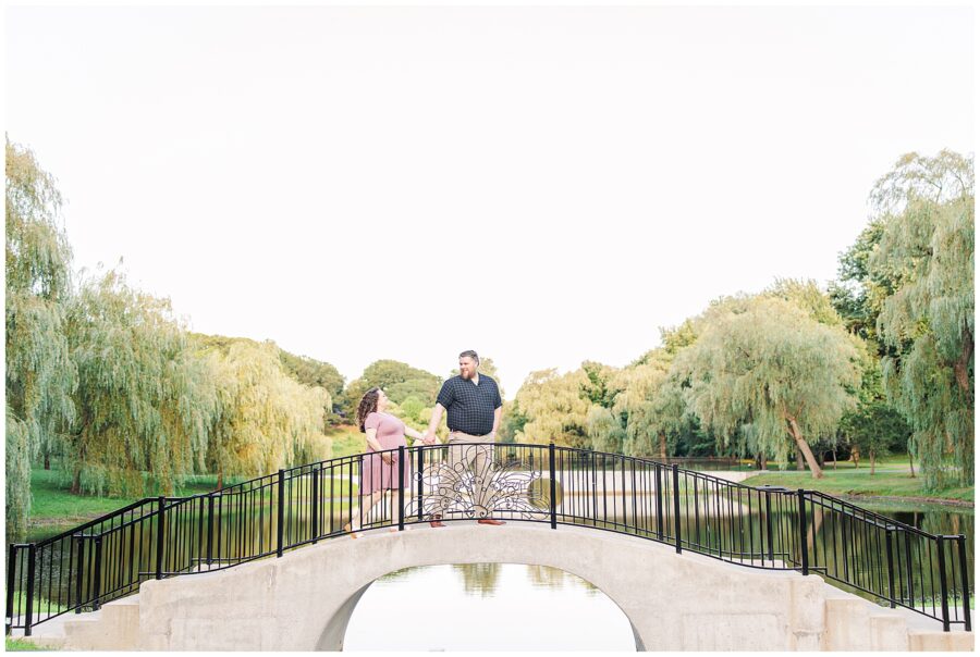 A couple holding hands while standing on a small arched bridge over a pond at Larz Anderson Park, surrounded by lush trees. The man looks at his pregnant partner as they enjoy the scenic surroundings. Perfect example of a Boston maternity photographer capturing a serene moment.