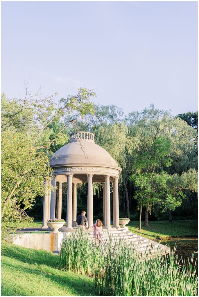 The couple walking toward a classic, dome-shaped pavilion in Larz Anderson Park. Tall grass and trees surround the structure, creating a peaceful setting for Boston maternity photographer sessions.