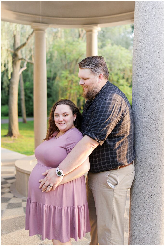 The couple standing close together at the pavilion in Larz Anderson Park. The man stands behind the pregnant woman, his hand resting on her belly, while they smile at the camera.