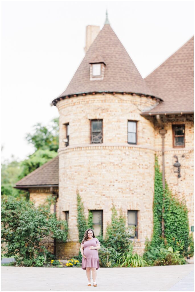 A pregnant woman stands in front of a historic stone building with ivy climbing the walls at Larz Anderson Park in Brookline, MA. This serene location is ideal for a Boston maternity photographer to capture timeless moments.