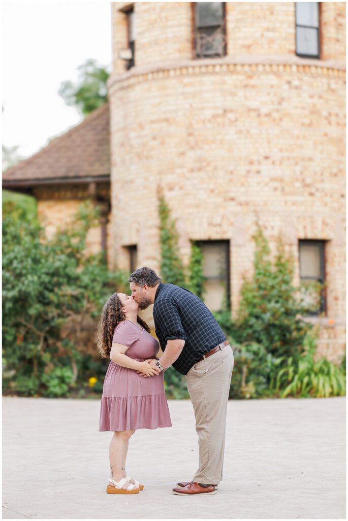 The couple shares a kiss in front of a large, round stone turret at Larz Anderson Park. The picturesque setting showcases the beauty of the location, making it a popular choice for a Boston maternity photographer.