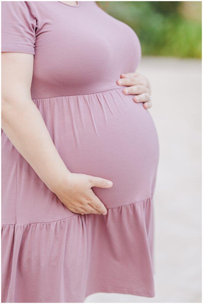 Close-up of a pregnant woman’s belly, cradled gently by her hands. She wears a lavender dress, and the soft background complements the intimate moment.