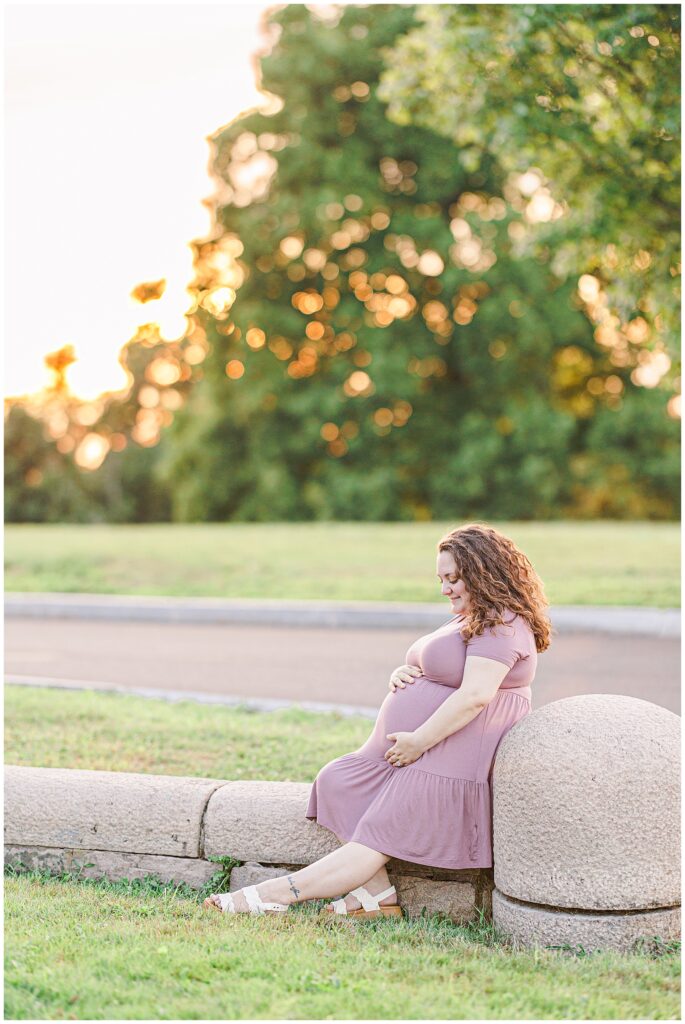 A pregnant woman sits on a stone barrier, cradling her belly, as the sun sets behind her. The warm light and natural surroundings make this a perfect scene for a Boston maternity photographer.