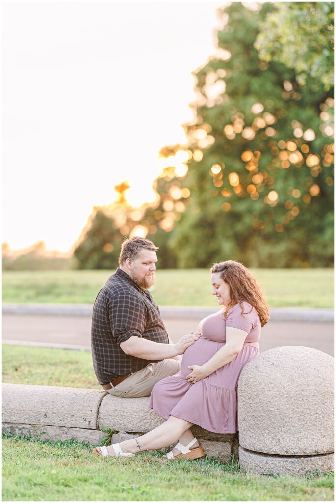 The couple sits together on a stone barrier at sunset in Larz Anderson Park. The man places his hand on the woman’s belly as they share a tender moment, ideal for Boston maternity photography.