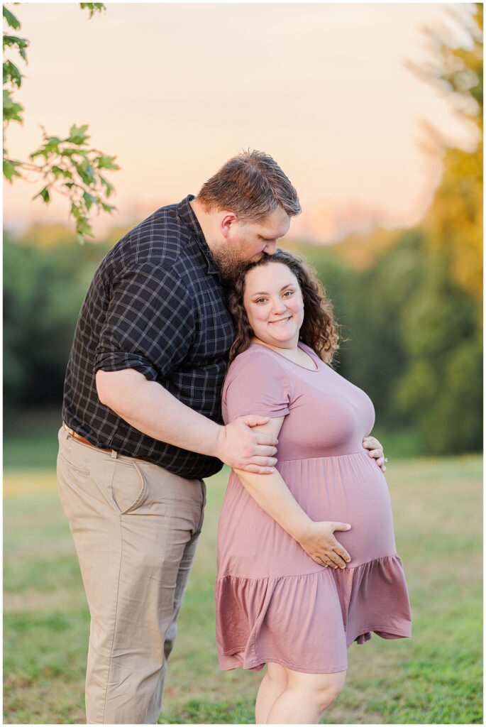 The man kisses the top of the pregnant woman’s head as they stand together in a grassy field, bathed in the soft glow of the evening light at Larz Anderson Park.