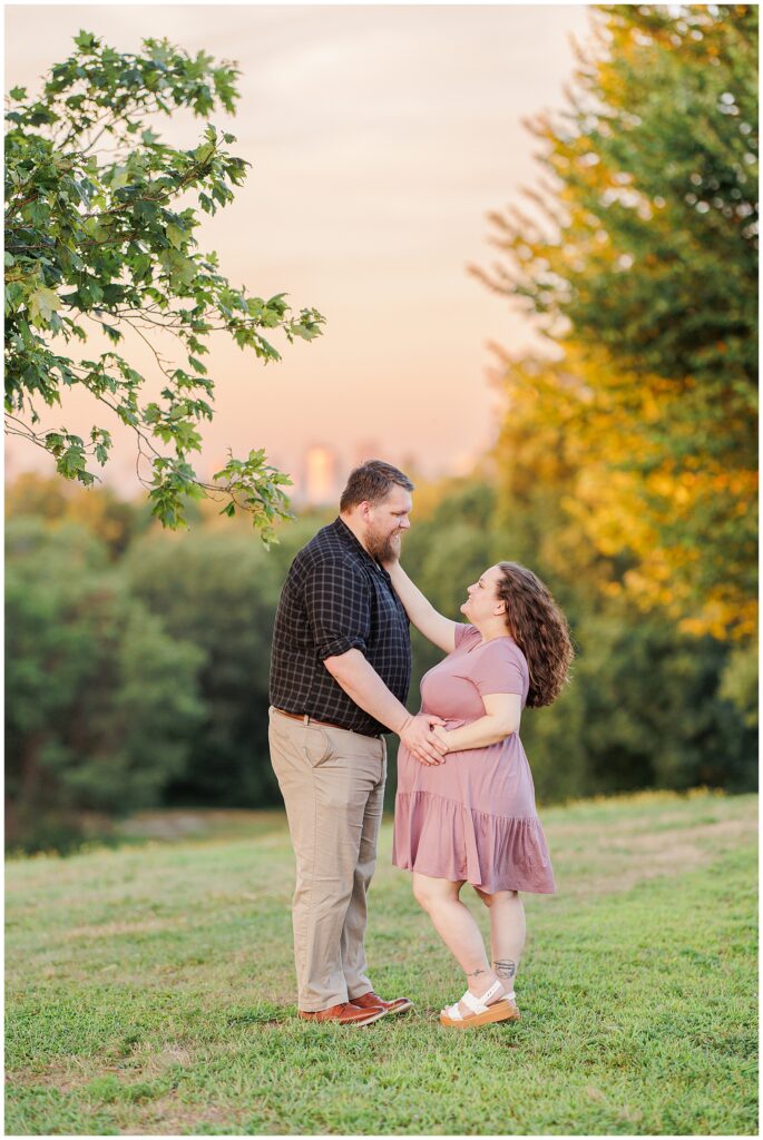 The couple stands facing each other, holding hands in a grassy area of Larz Anderson Park. The backdrop of sunset and greenery enhances the peaceful atmosphere, a wonderful scene for a Boston maternity photographer