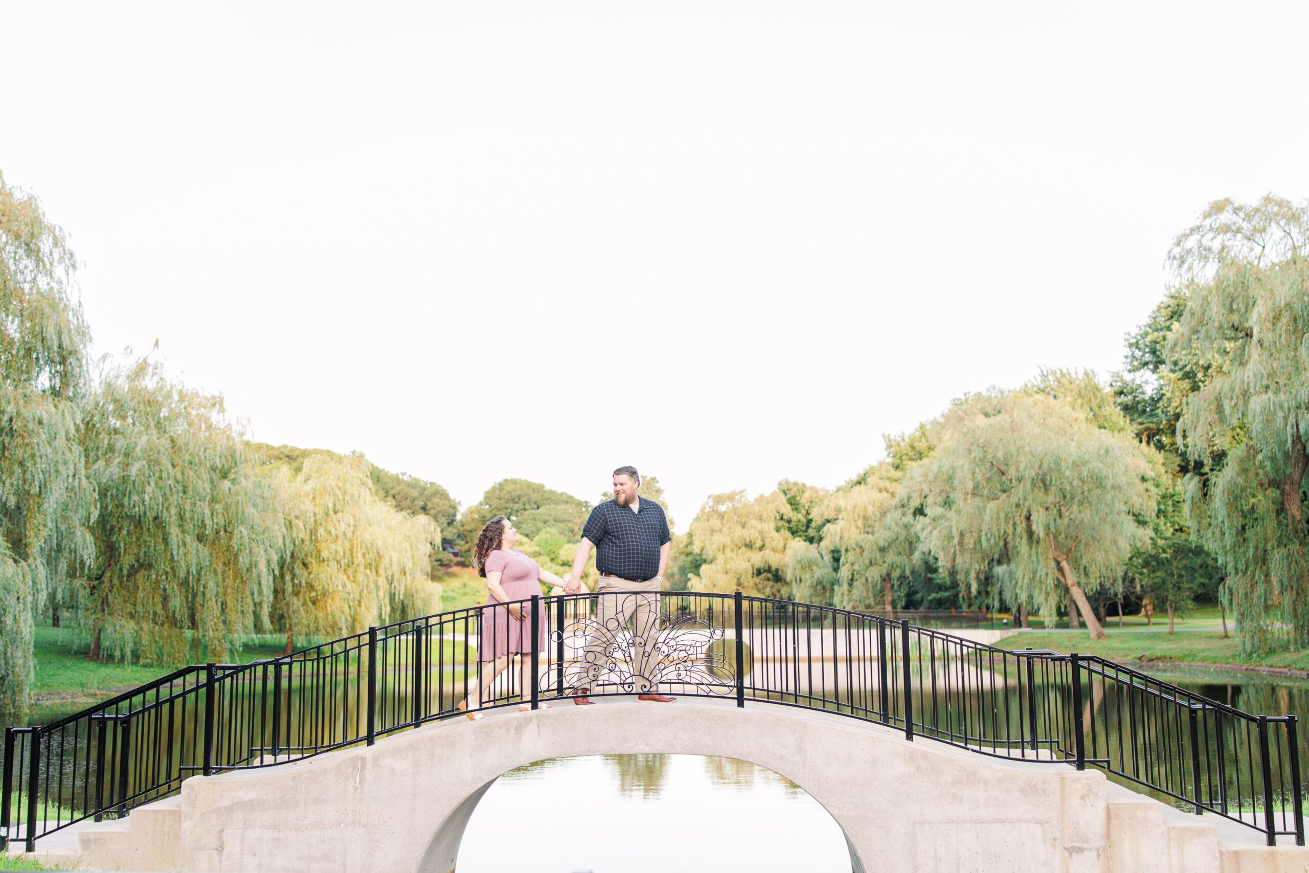 The couple holds hands on a small arched bridge over a pond at Larz Anderson Park, with lush willow trees in the background. This peaceful scene is often sought out by Boston maternity photographers for its scenic beauty.