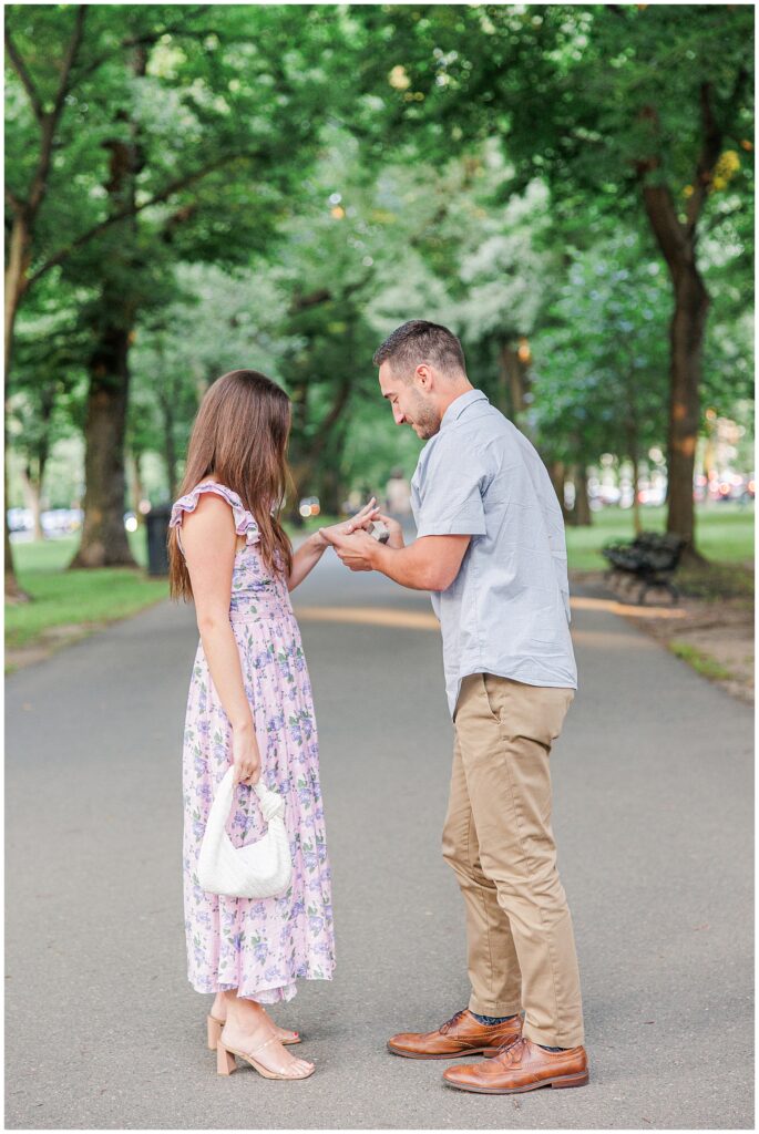 The man places a ring on the woman’s finger as they stand together on the park path. The woman smiles and holds a small white bag, with green trees and benches surrounding them at Commonwealth Avenue Mall.