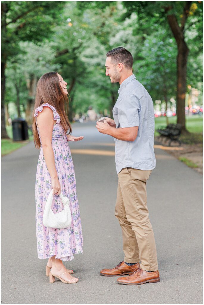 The couple laughs together after the proposal, standing face-to-face on the pathway at Commonwealth Avenue Mall. The woman is smiling, holding a small white bag, while the man holds the ring box.