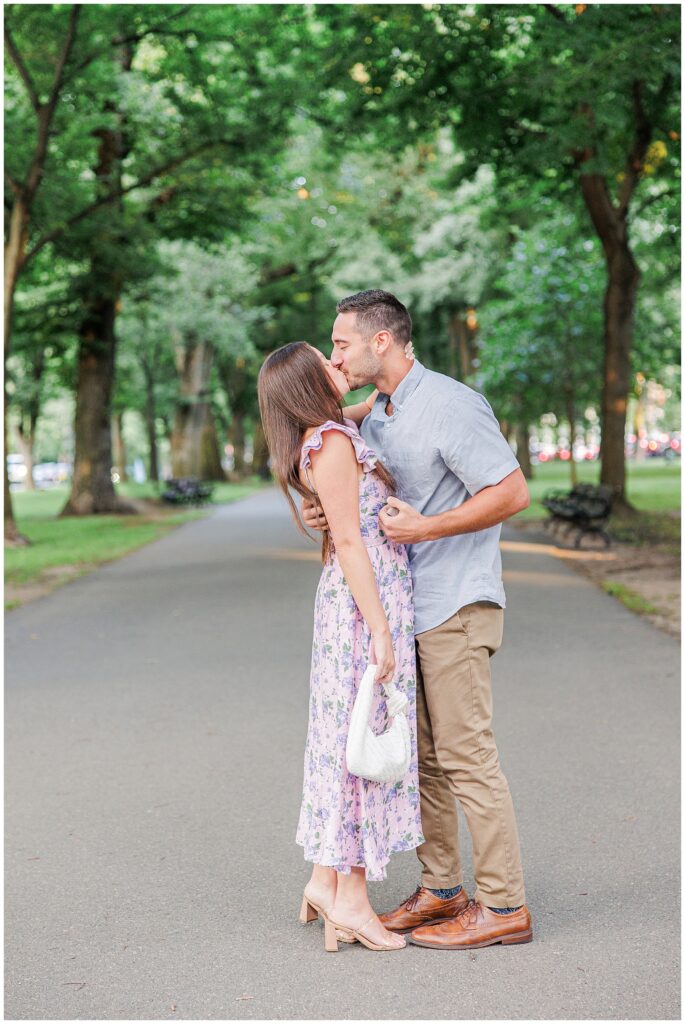 The couple shares a kiss after the proposal, standing close on the park pathway. The man holds the woman gently, with the tree-lined Commonwealth Avenue Mall as the backdrop.