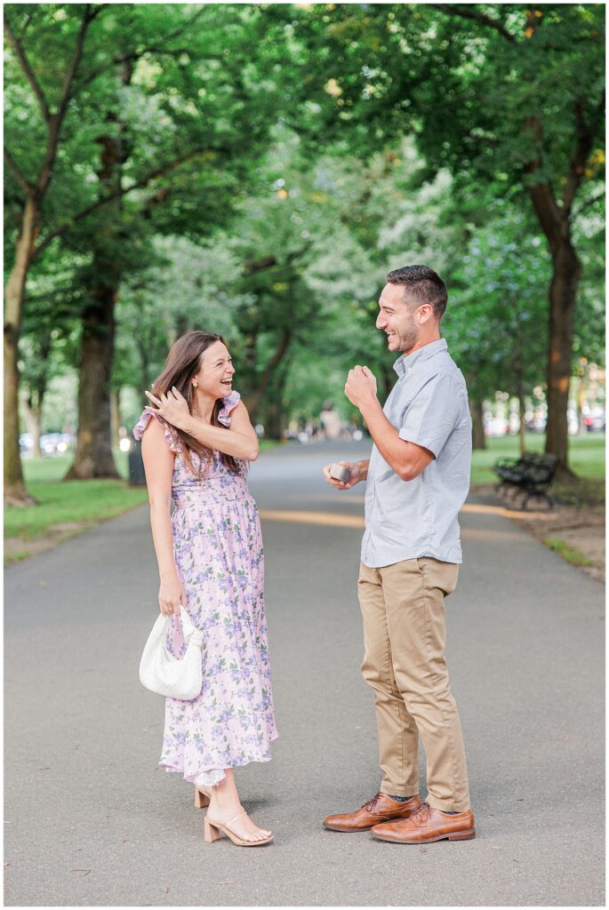 The couple stands smiling and talking after the proposal. The man holds the ring box, and the woman holds her small white bag, with the tree-filled park behind them on Commonwealth Avenue Mall.