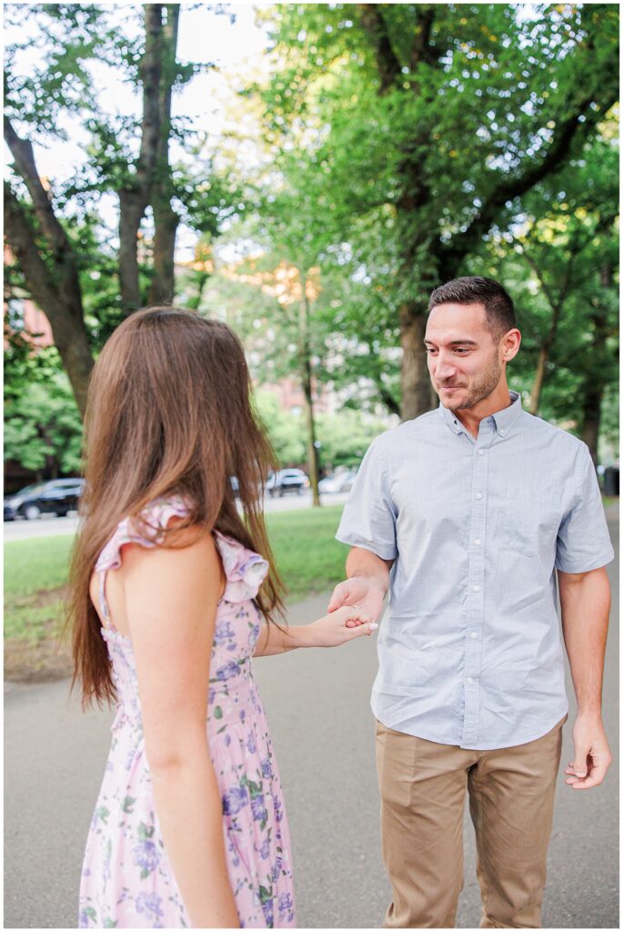 A close-up image of a man and woman standing on a tree-lined path at Commonwealth Avenue Mall in Boston. The man, wearing a light blue shirt and khaki pants, holds the woman’s hand and smiles. The woman, dressed in a floral lavender dress, has her back to the camera. Green trees and parked cars are visible in the background.