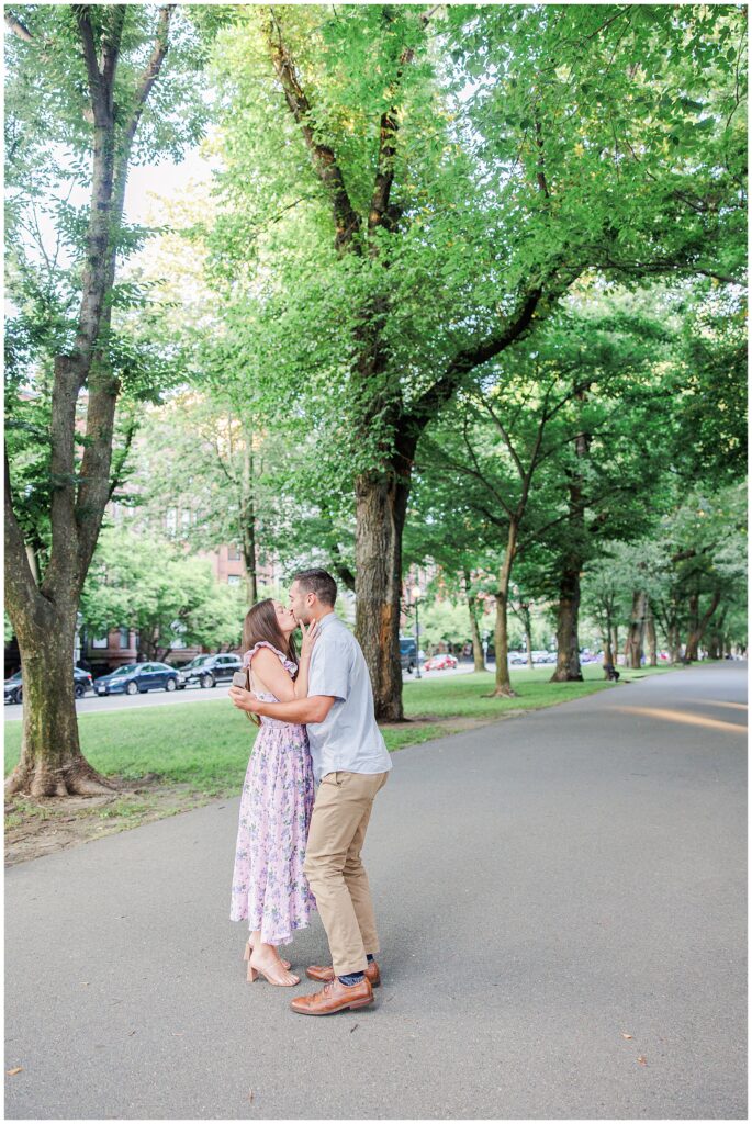 The couple embraces and kisses on the pathway, with large trees surrounding them. The peaceful greenery of Commonwealth Avenue Mall adds to the romantic setting.