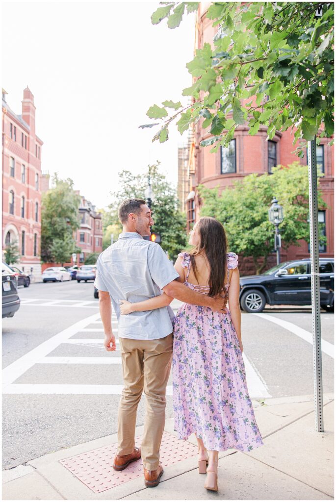 The couple walks away arm-in-arm down a city street near Commonwealth Avenue Mall, with the man wearing a light blue shirt and the woman in a floral dress. Brick buildings and parked cars are visible as they walk away from the proposal.