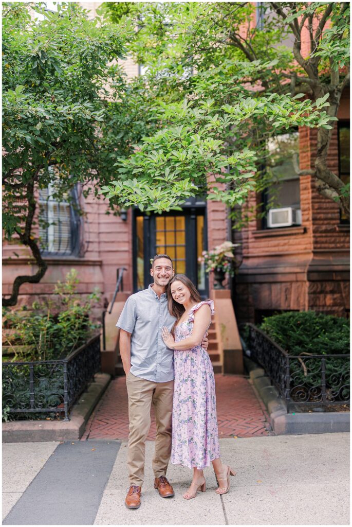 A couple stands together, smiling and embracing in front of a brick building on Commonwealth Avenue in Boston. The man is wearing a light blue shirt and khaki pants, and the woman is in a floral lavender dress. Green trees and plants surround the entrance to the building.