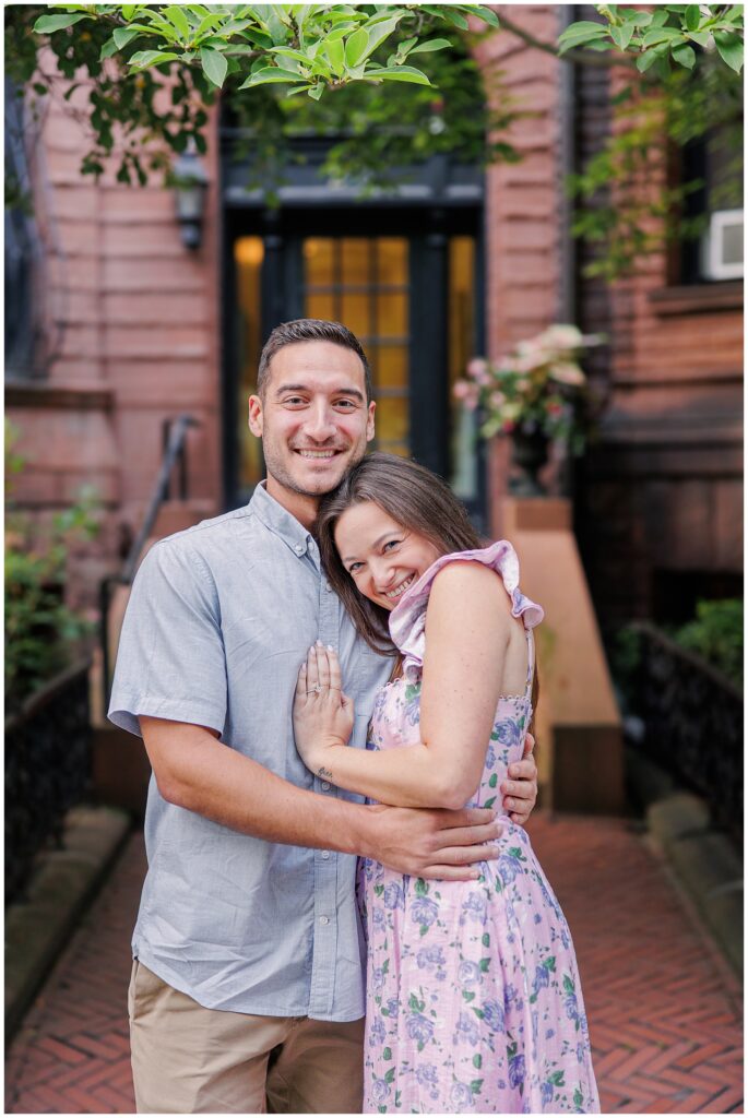 A close-up of the couple smiling and holding each other in front of a brick building on Commonwealth Avenue. The man has his arm around the woman, and she rests her head on his chest, looking happy.