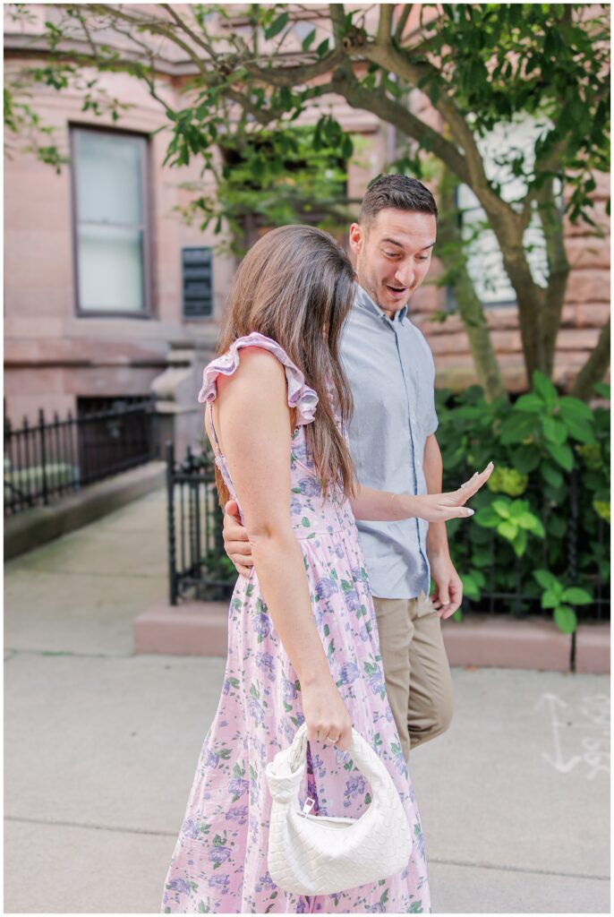 The couple walks on the sidewalk, and the woman shows off her engagement ring. The man, smiling, has his arm around her as they pass a tree in front of a historic brick building on Commonwealth Avenue.