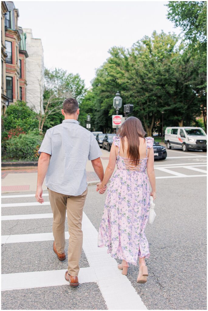 The couple is seen from behind, holding hands and walking across a crosswalk on Commonwealth Avenue. The woman wears a floral lavender dress, and the man is in a light blue shirt and khaki pants. Brick buildings and parked cars are visible along the street.