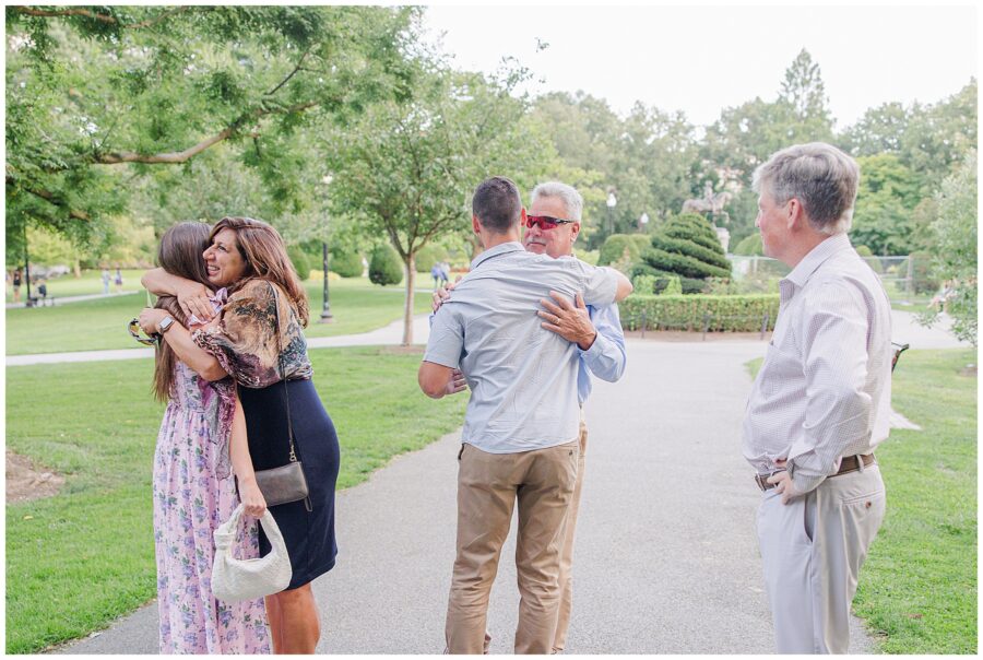 A couple embraces their family after the proposal in Boston Public Garden. The woman hugs another woman, while the man hugs an older man wearing sunglasses. A man stands nearby, watching the happy moment.