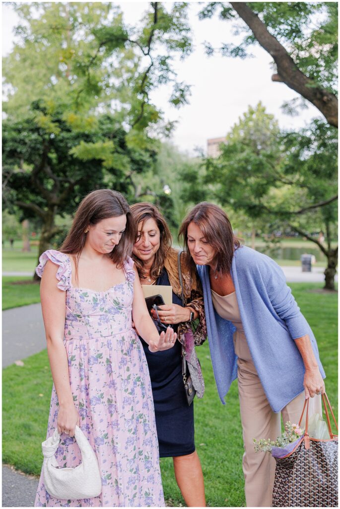 The woman, wearing a floral lavender dress, shows off her engagement ring to two women who smile and lean in to get a closer look. They are standing in the Boston Public Garden, surrounded by trees and greenery.