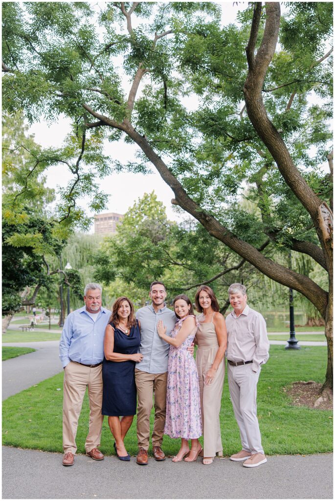 he couple stands with their family in Boston Public Garden, posing for a group photo. The couple is in the center, smiling and holding each other, with their family members standing on either side.