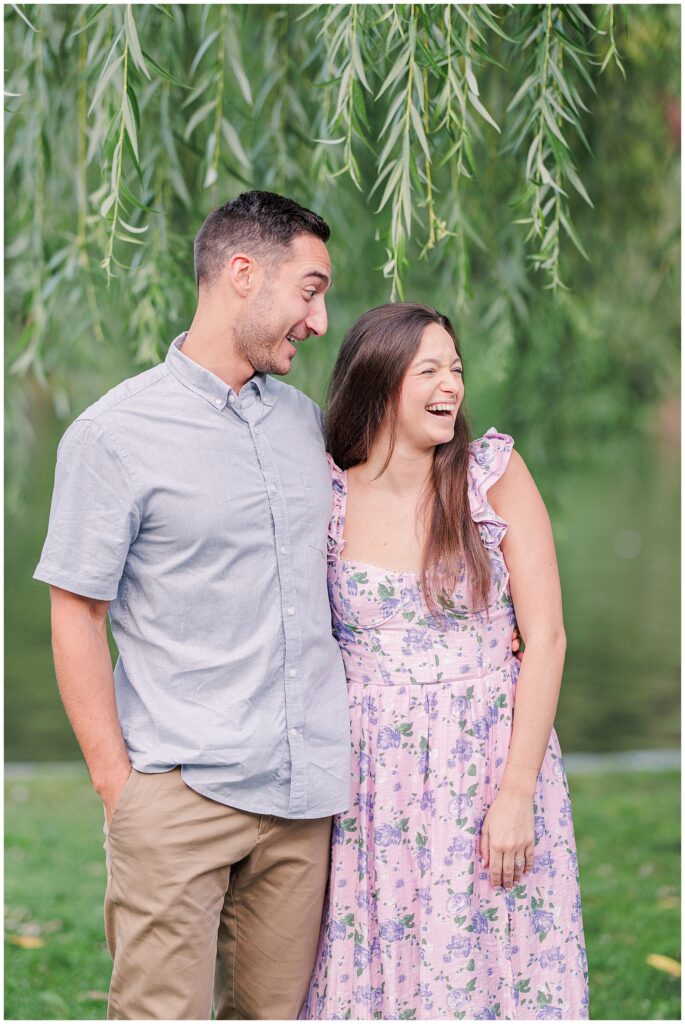 The couple stands close together under a willow tree in Boston Public Garden, both smiling and looking off to the side. The greenery and pond in the background create a peaceful, romantic atmosphere.