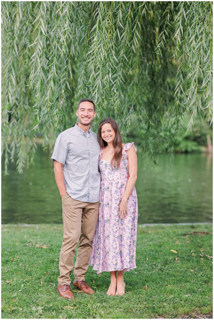 The couple stands in front of a willow tree, smiling directly at the camera in Boston Public Garden. The woman wears a floral lavender dress, and the man wears a light blue shirt and khaki pants.