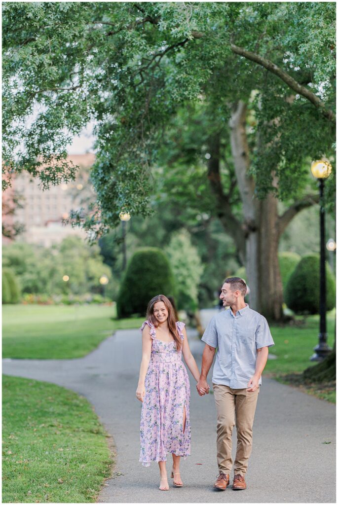 The couple walks hand-in-hand along a pathway in Boston Public Garden, smiling at each other. The lush green trees and grass surrounding them create a serene backdrop.