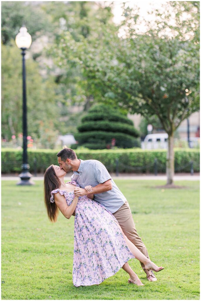 The man dips the woman and kisses her on a grassy area in Boston Public Garden. The woman holds onto his arm, smiling, as trees and lamp posts surround them.