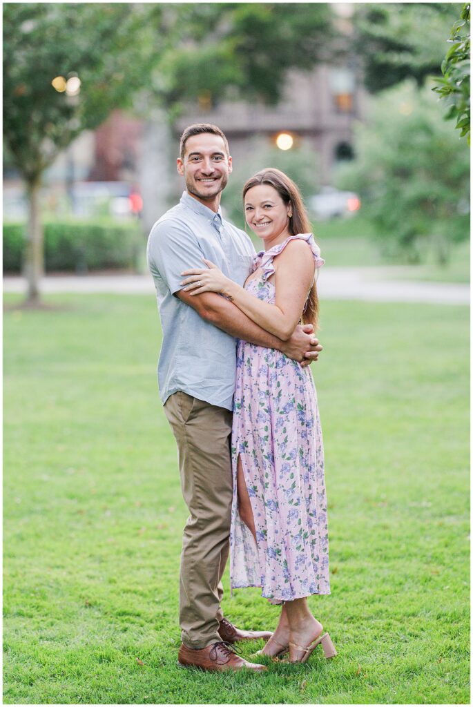 The couple poses, standing close together on the lawn in Boston Public Garden, smiling at the camera. The woman rests her hand on the man’s chest, showing her engagement ring.