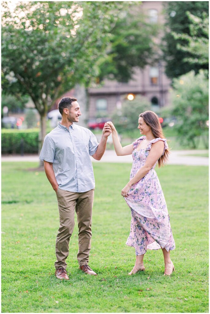 The couple holds hands and smiles at each other as they stand on the lawn in Boston Public Garden. The woman’s floral dress flows in the breeze as they enjoy a joyful moment outdoors.