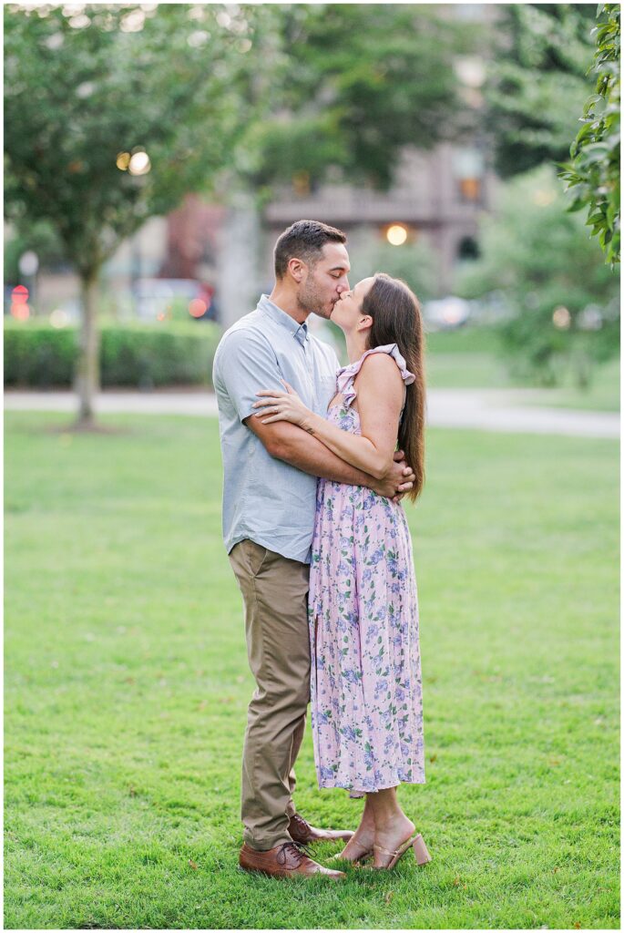 The couple shares a kiss in Boston Public Garden, standing in a grassy area with trees in the background. They embrace each other, creating a tender moment in the park.