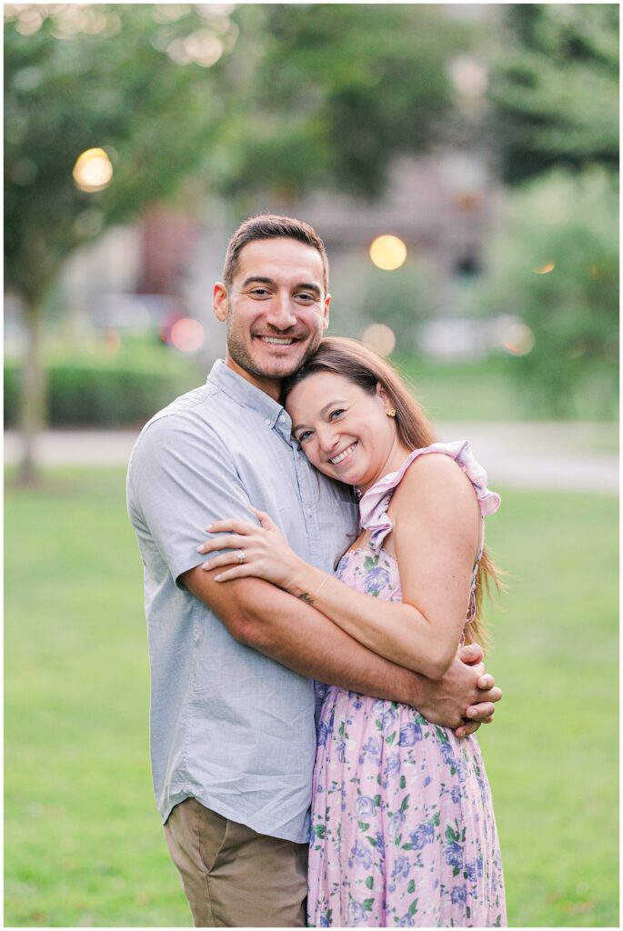 A couple hugs in Boston Public Garden. The man, in a light blue shirt, holds the woman, who is wearing a floral lavender dress. She looks at the camera, both holding each other closely, with a soft green park background.