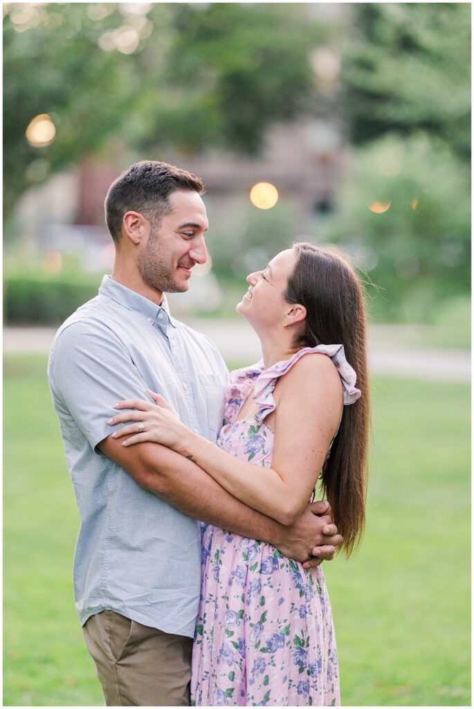 A couple embraces in Boston Public Garden. The man, in a light blue shirt, looks down smiling at the woman, who is wearing a floral lavender dress. She gazes up at him, both holding each other closely, with a soft green park background.