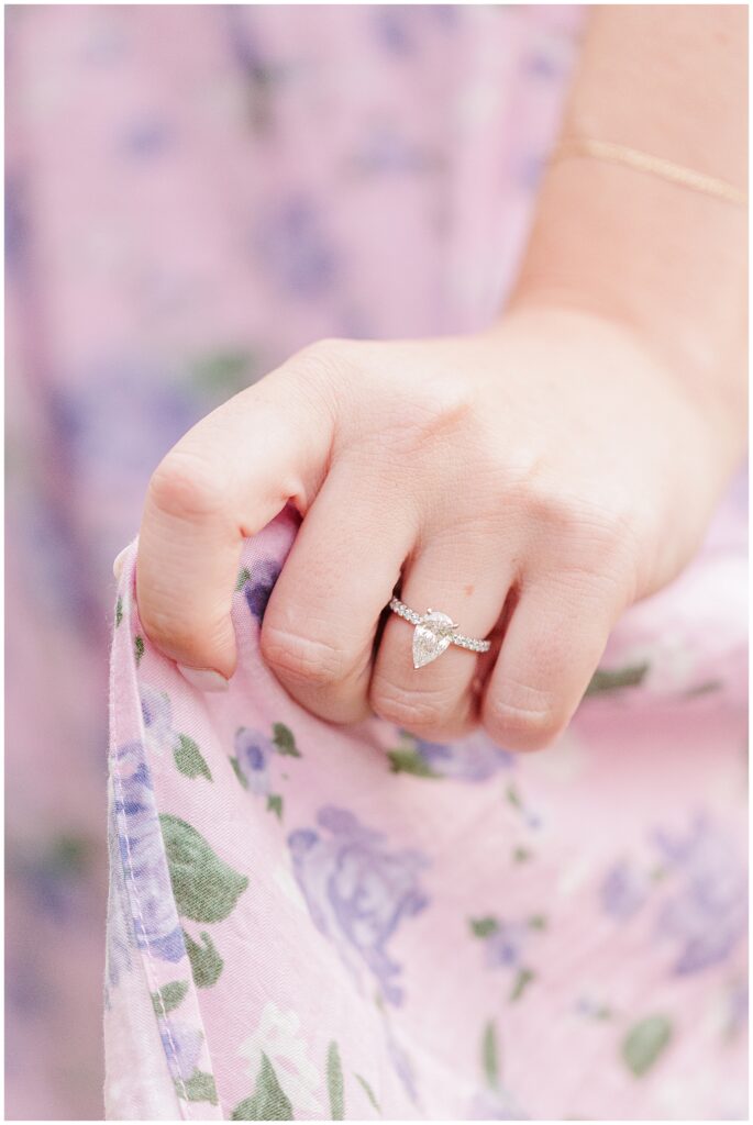 A close-up of the woman’s hand holding her floral dress, showing her pear-shaped diamond engagement ring with a sparkling band.